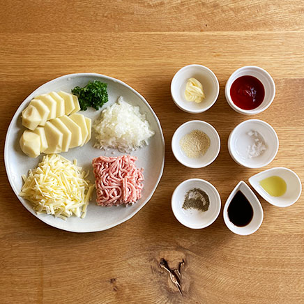 丸型グラタン皿1皿分のじゃがいものミートソースグラタンに使用した材料(じゃがいも、豚ひき肉、玉ねぎ、パセリ、プロセスチーズ、バター、ウスターソース、ケチャップ、コンソメ、塩、黒こしょう、オリーブオイル)